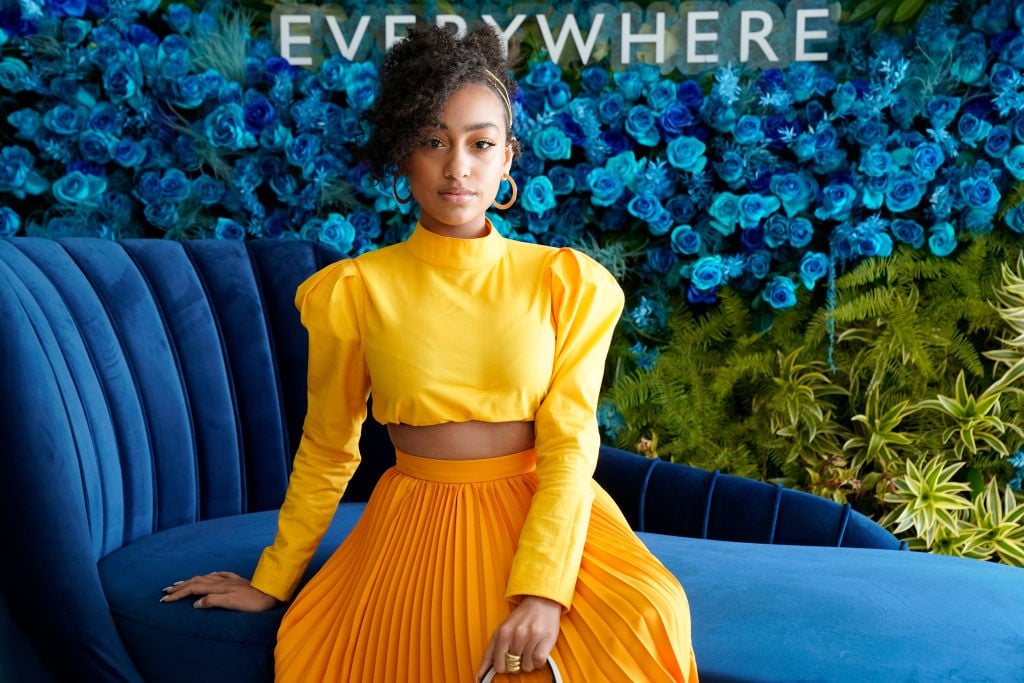 Lexi Underwood wearing a gold yellow outfit on a dark teal couch in front of a teal and green backdrop