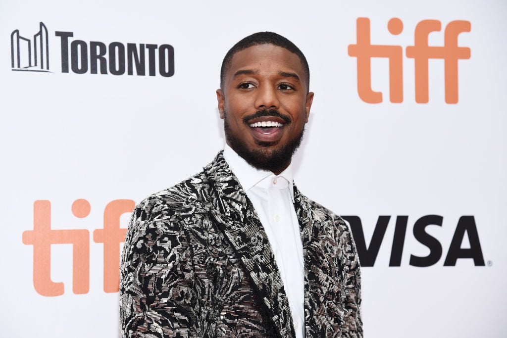 Michael B. Jordan smiling, looking away from the camera, in front of a white background with repeating logos