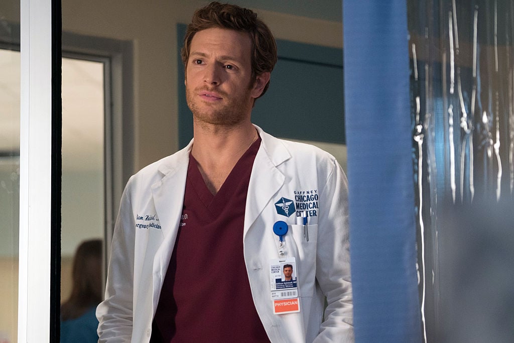 Nick Gehlfuss as Will Halstead in a doctor's coat and dark red scrubs