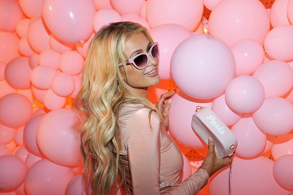 Paris Hilton smiling wearing sunglasses in front of a wall of pink balloons