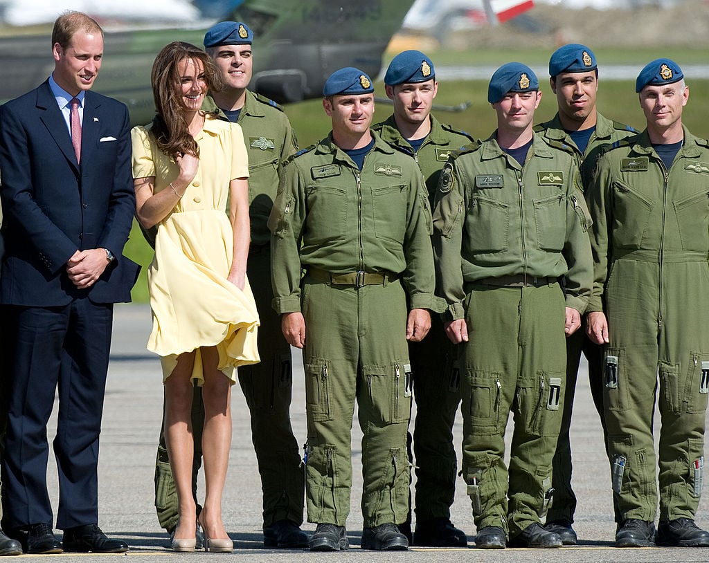 Kate Middleton keeps her hair and dress from blowing in the wind standing next to Prince William and soldiers