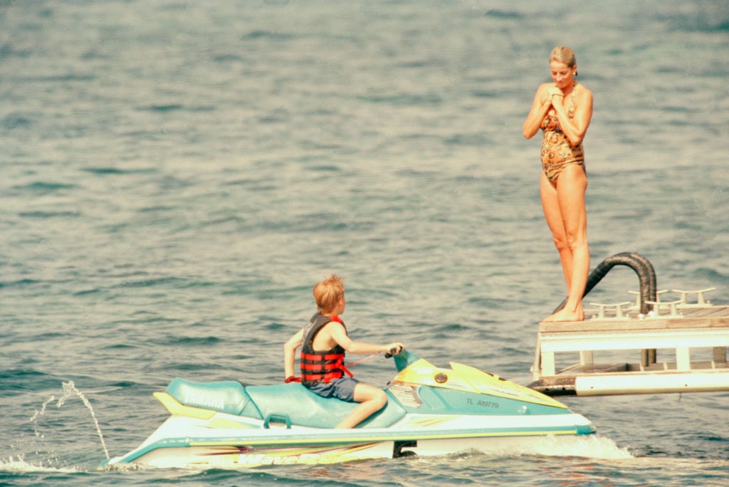 Princess Diana stands on a dock while Prince Harry sits on a jet ski, 1997