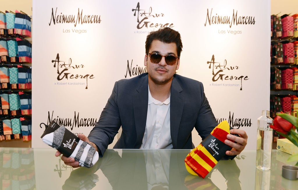 Rob Kardashian wearing sunglasses and a gray blazer holding up two pairs of socks