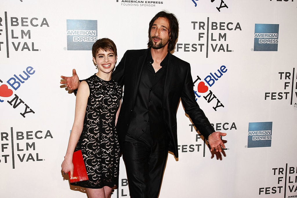 Sami Gayle and Adrien Brody smiling in front of a repeating background