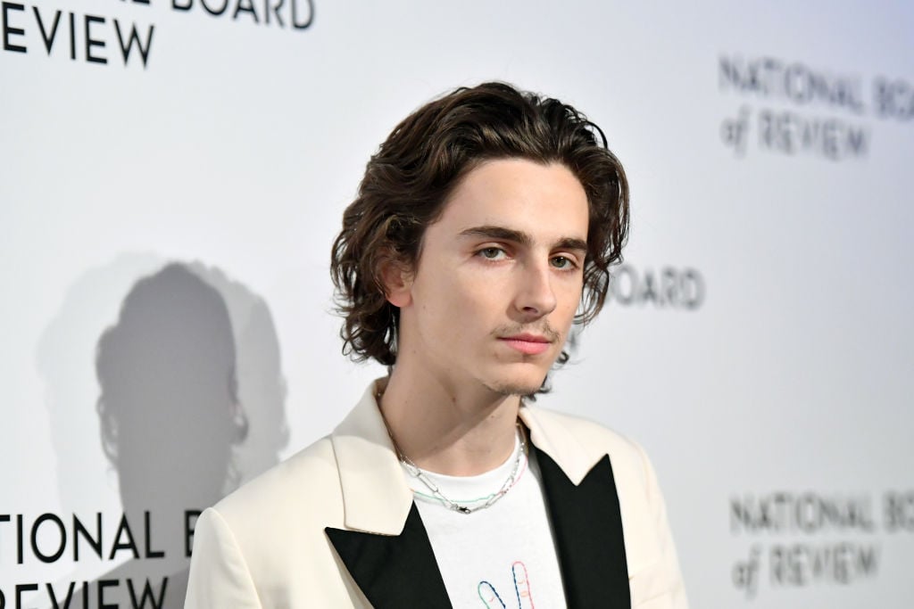 Timothée Chalamet smiling slightly to the side in front of a white background with a repeating logo