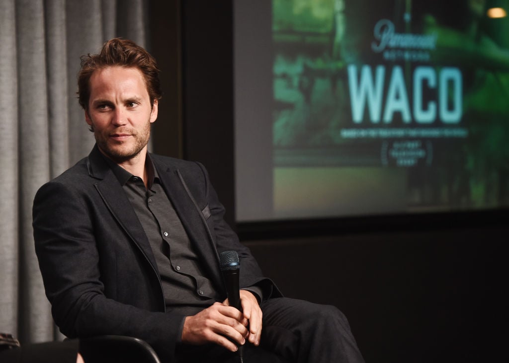 Taylor Kitsch seated holding a microphone in front of a 'Waco' screen