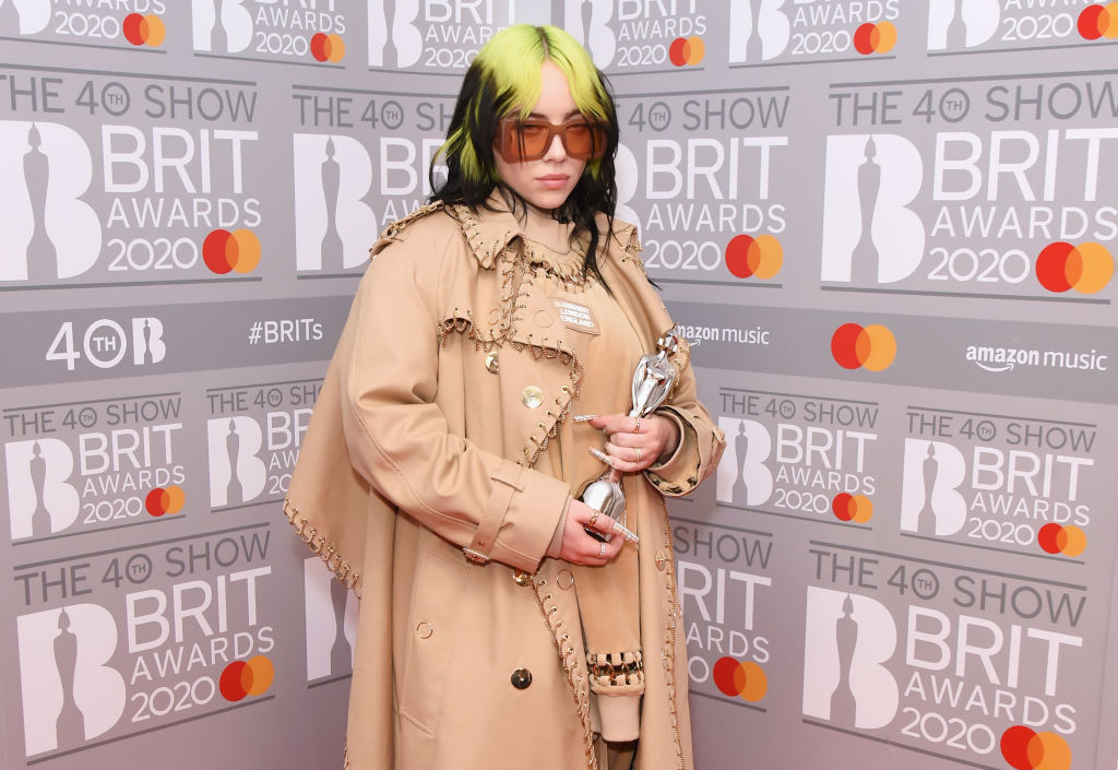 Billie Eilish wearing sunglasses, holding an award in front of a repeating background
