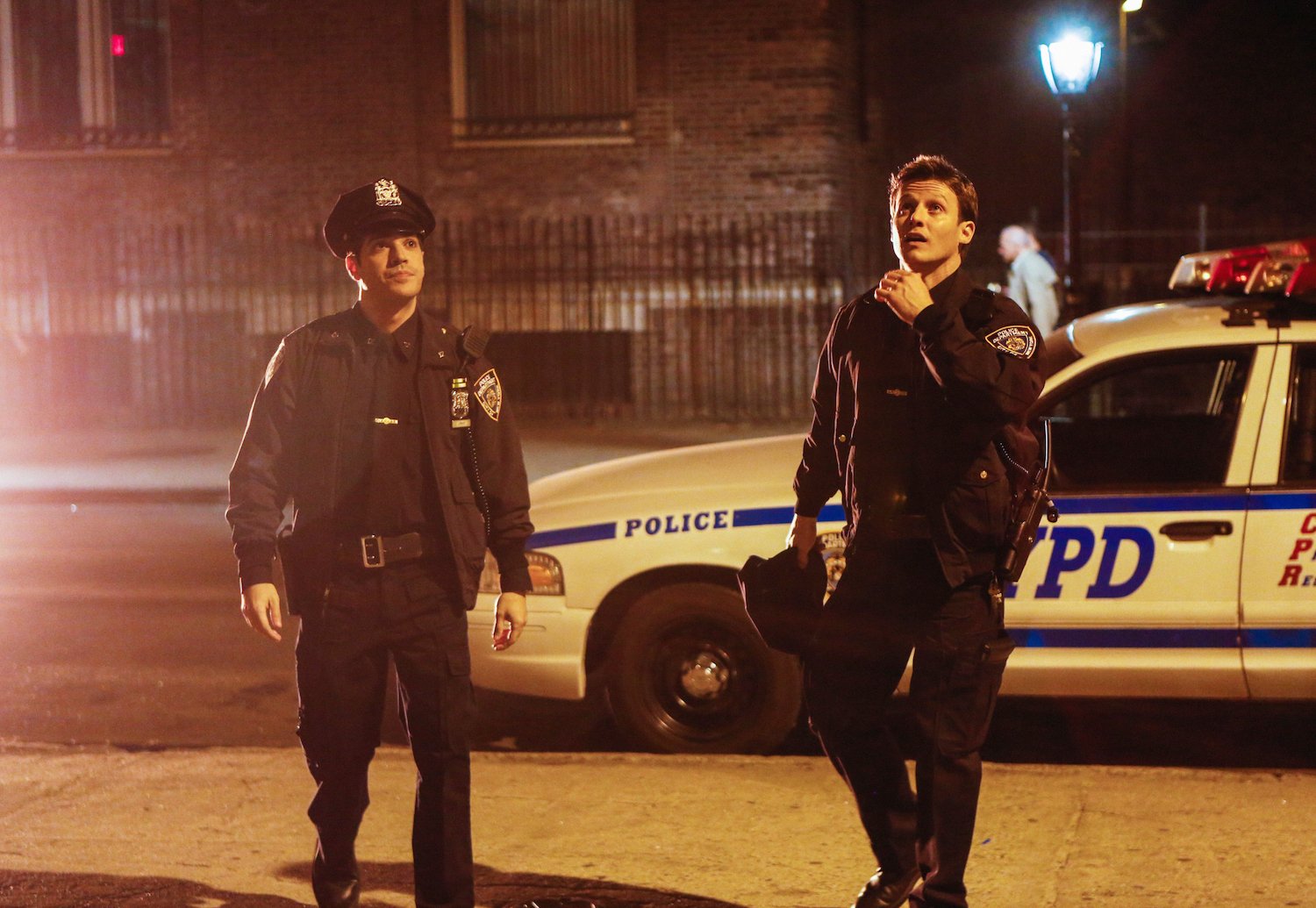 Vinny (left, Sebastian Sozzi) and Jamie (right, Will Estes) standing on the street in police uniform in front of an NYPD car