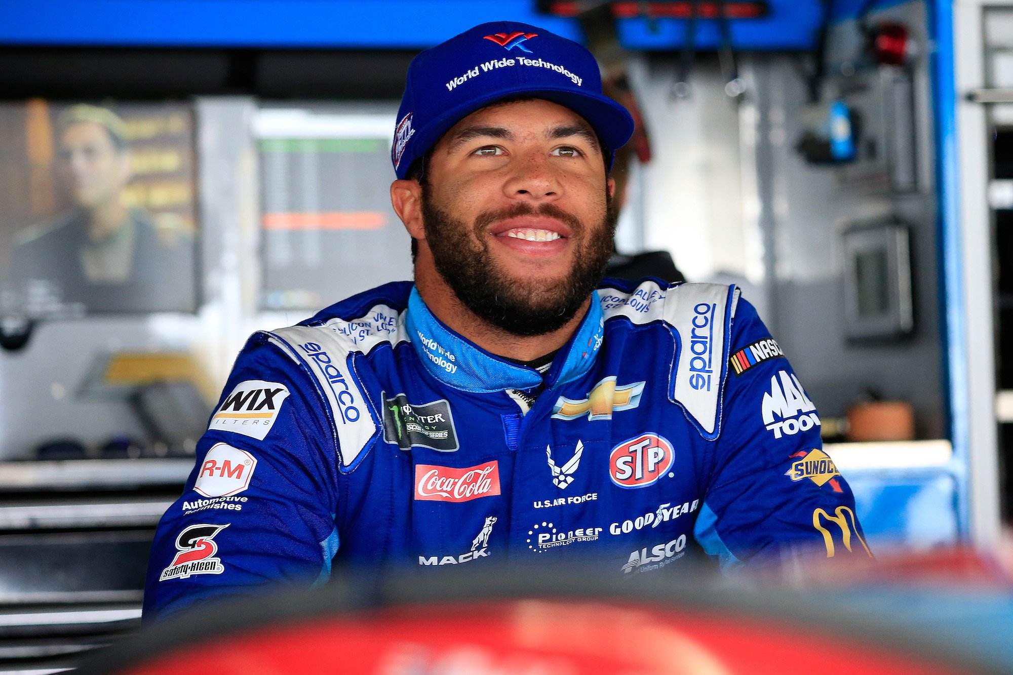 Bubba Wallace smiling in a racing jacket and blue hat, looking away from the camera