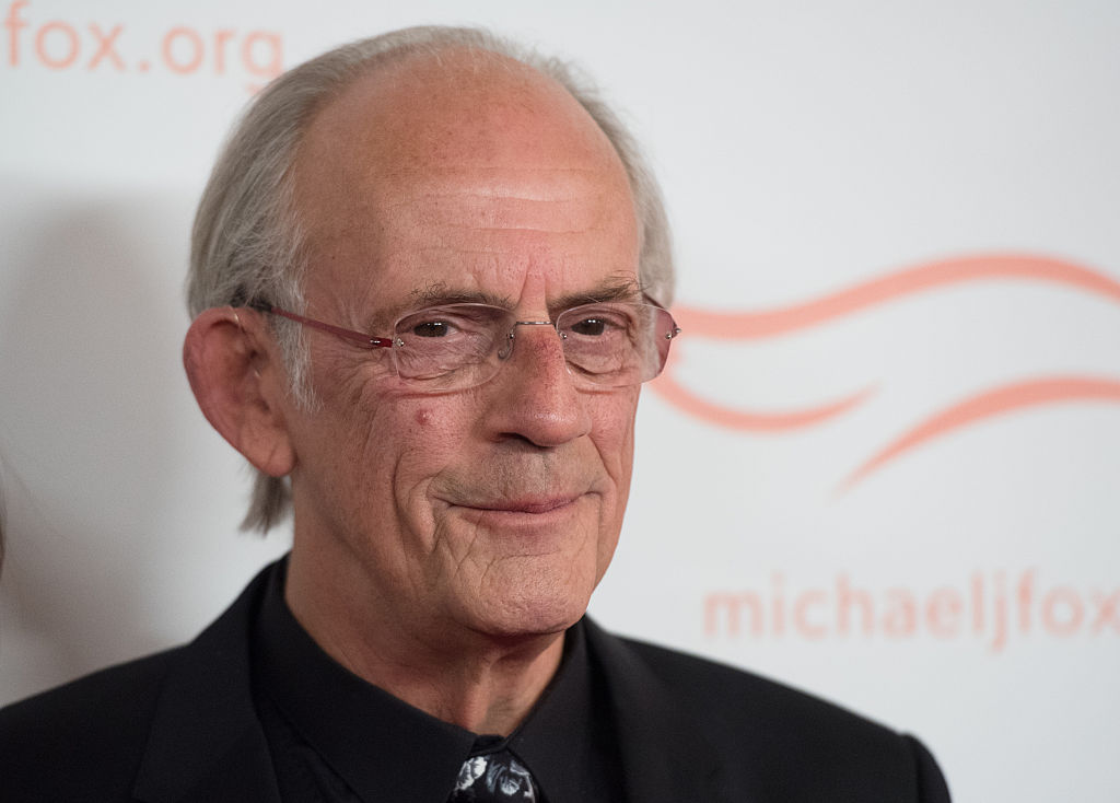 Christopher Lloyd smiling, looking away from the camera in front of a white background with a repeating logo