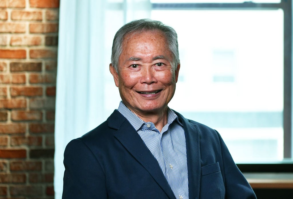 George Takei smiling in front of a window and brick wall
