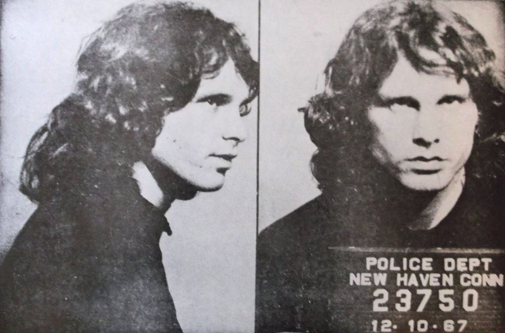 Jim Morrison, two photos in black and white side by side: first, profile mugshot; second, straight on mugshot holding arrest plaque with the date 12/7/67