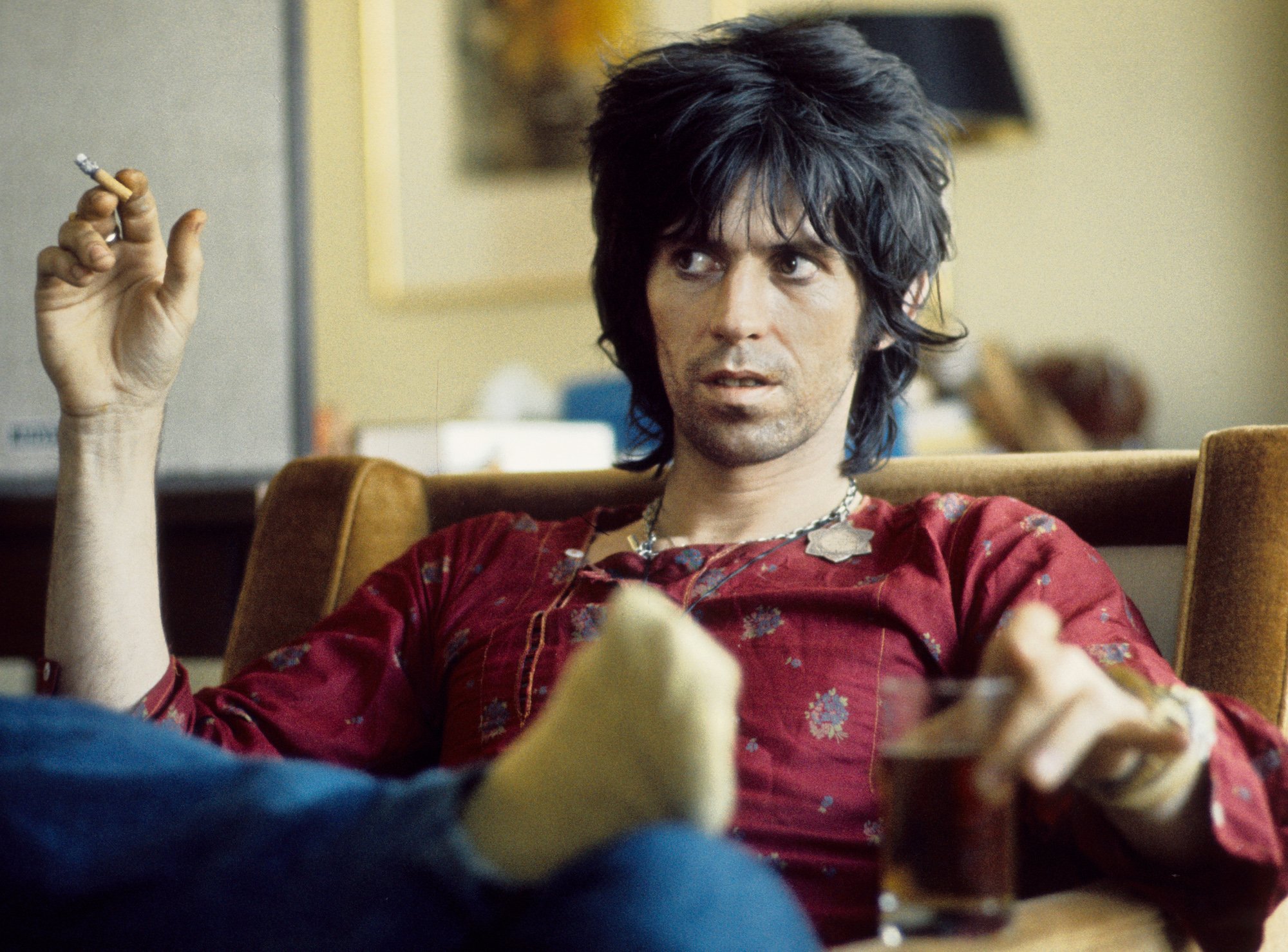 Keith Richards sitting in a chair, looking away from the camera, smoking and drinking