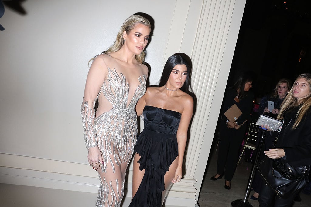 Khloé Kardashian and Kourtney Kardashian slightly smiling in front of a white wall