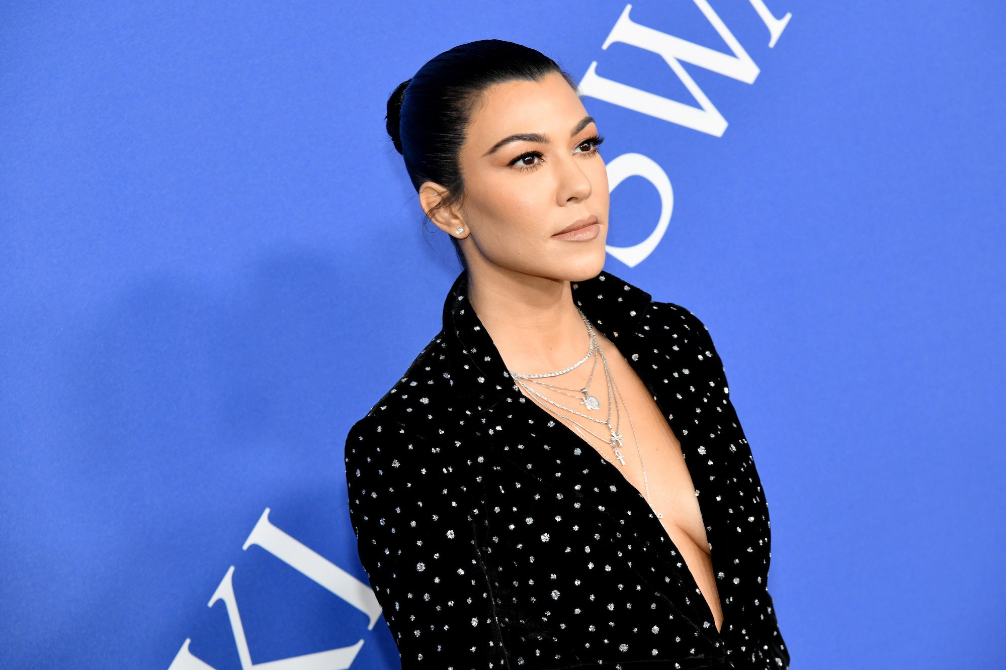 Kourtney Kardashian wearing a black blazer in front of a blue background, looking away from the camera