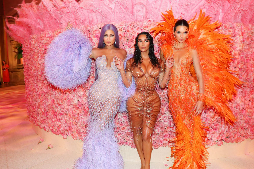 Kylie Jenner, Kim Kardashian West, and Kendall Jenner in purple, tan, and orange respectively, standing front of a pink background