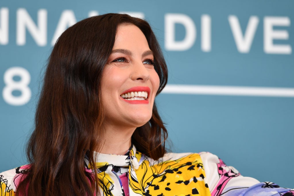 Liv Tyler smiling looking away from the camera in front of a blue background with a repeating white logo