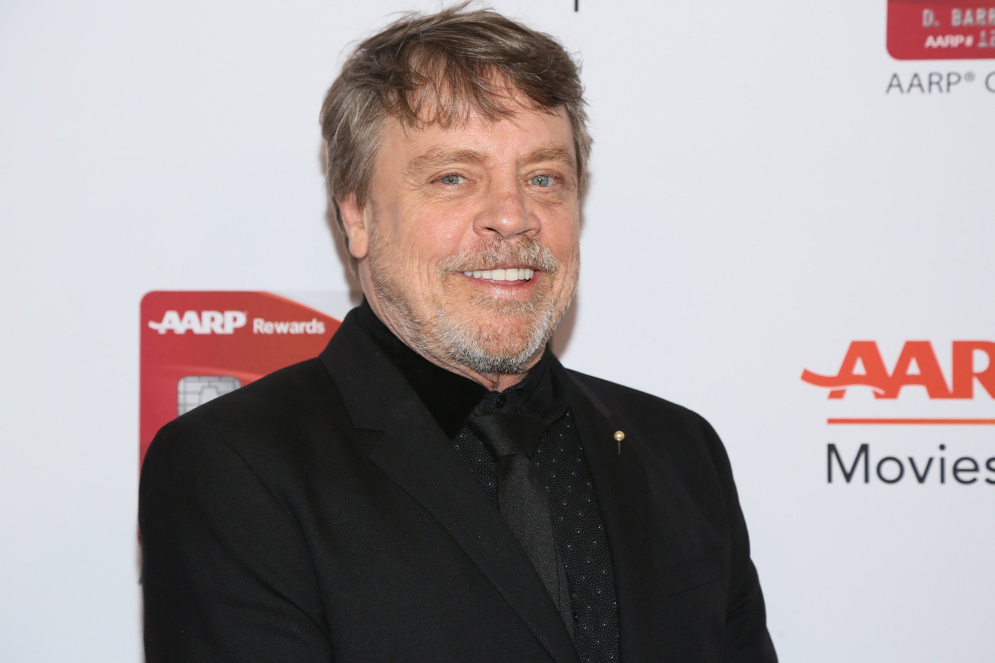 Mark Hamill smiling in front of a white background with a red repeating logo