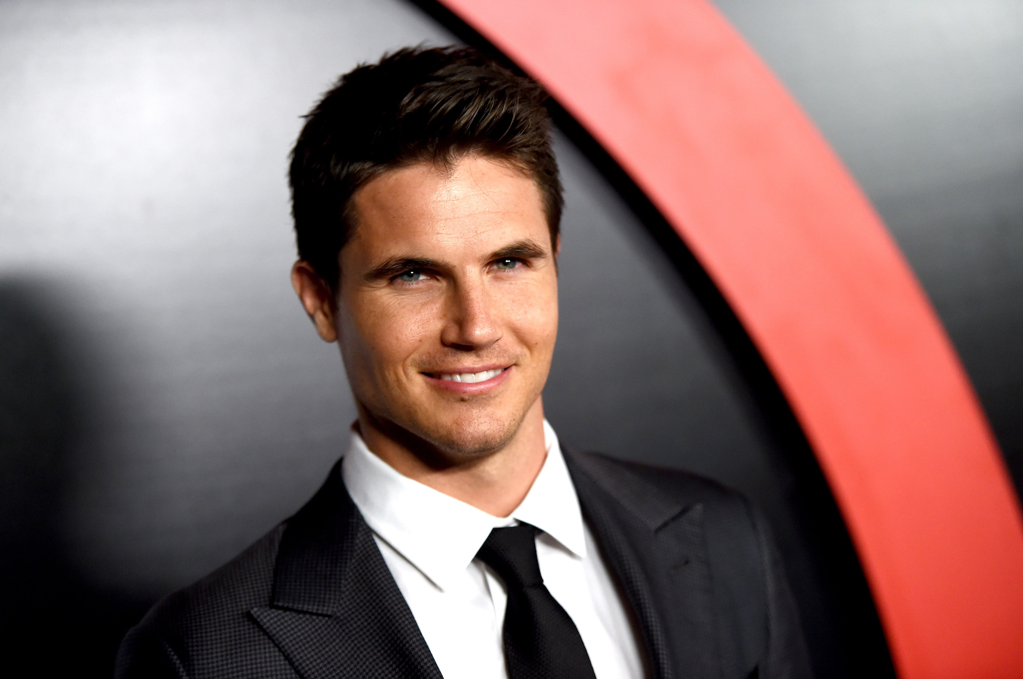 Robbie Amell smiling at the camera in front of a black and red background