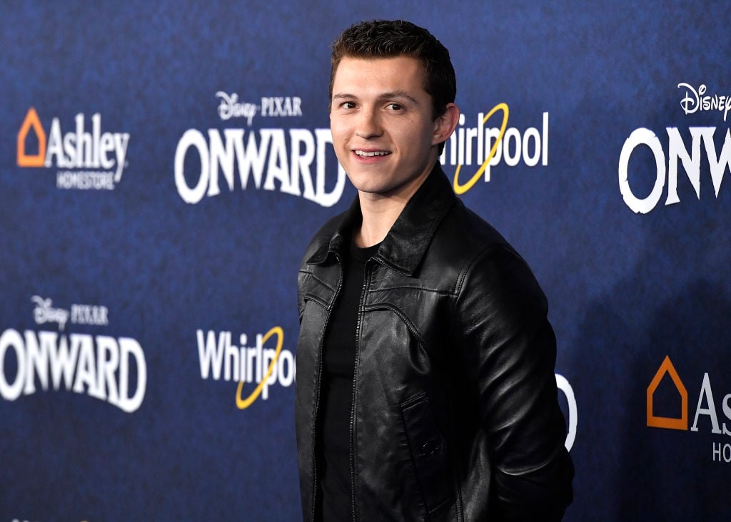 Tom Holland smiling in front of a dark blue background with a repeating logo