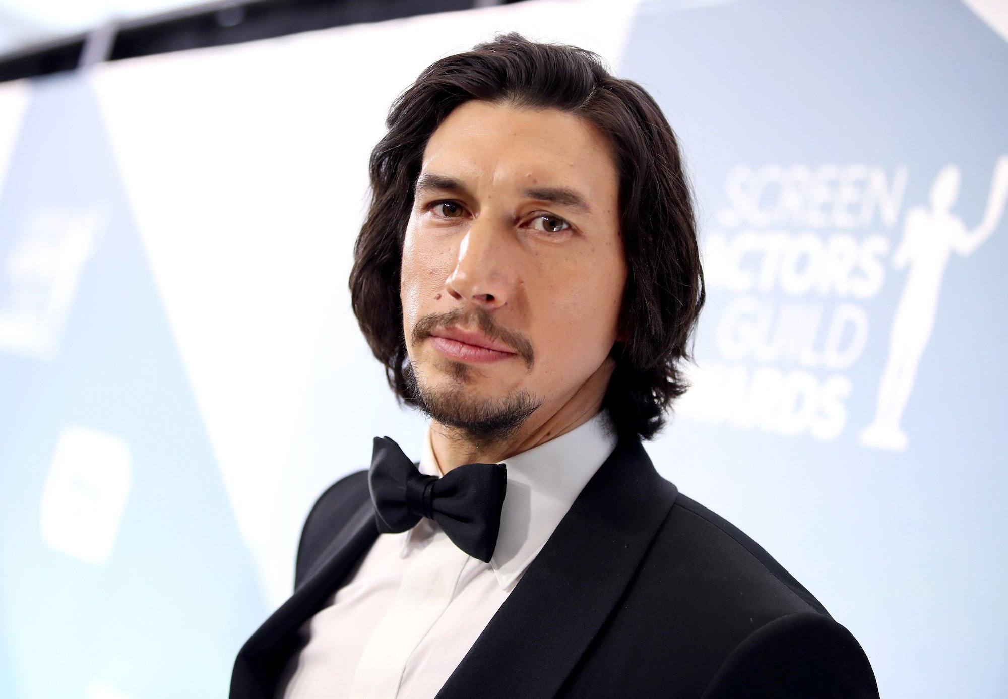 Adam Driver smiling standing in front of a white background