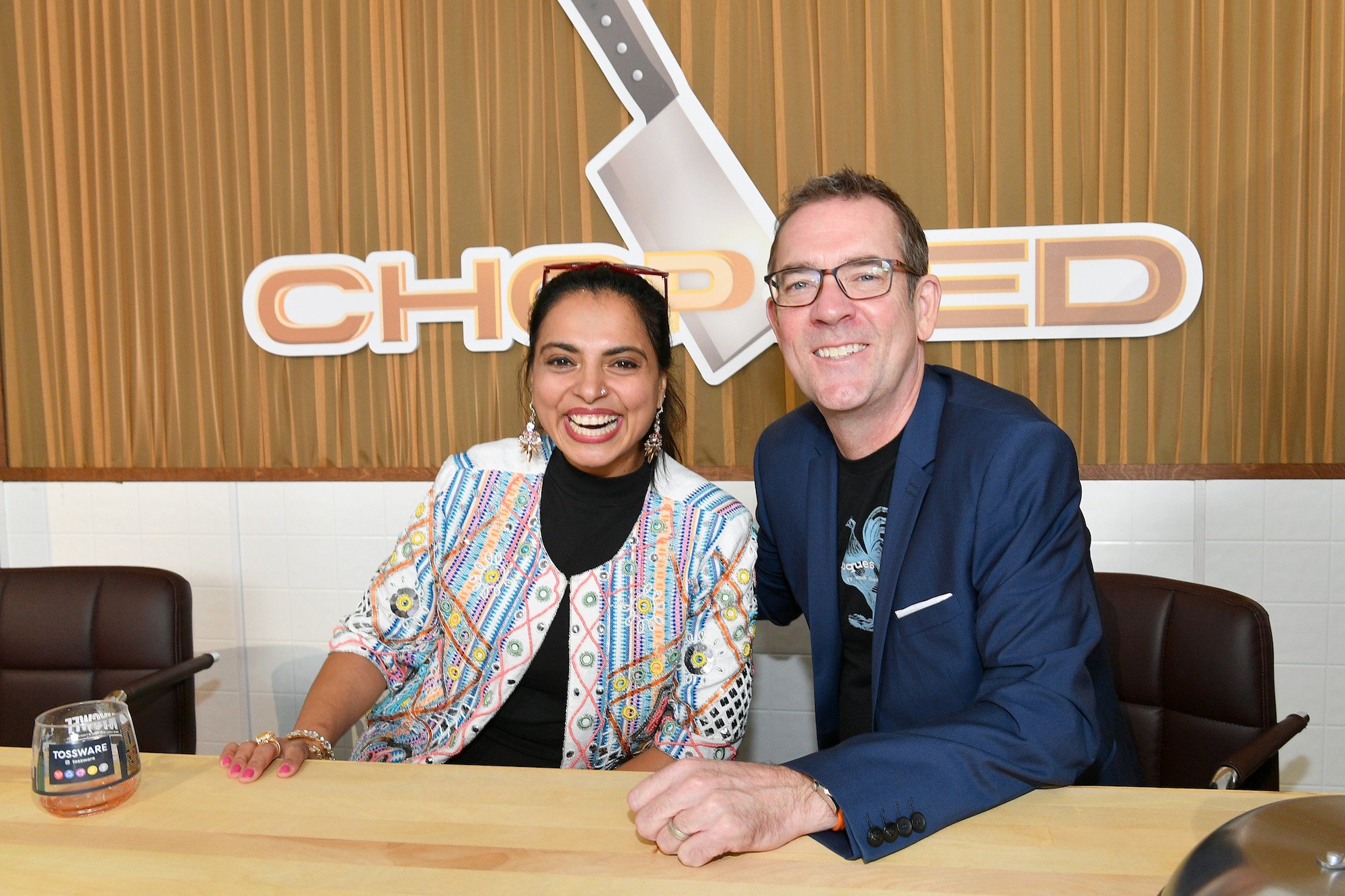 Maneet Chauhan and Ted Allen smiling in front of the Chopped logo