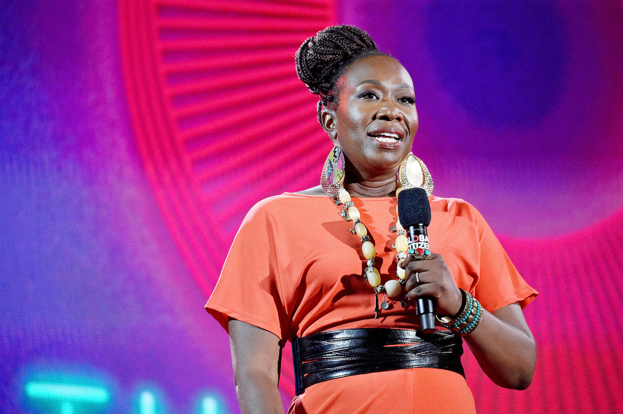 Joy Reid holding a microphone on stage in front of a pink and purple background