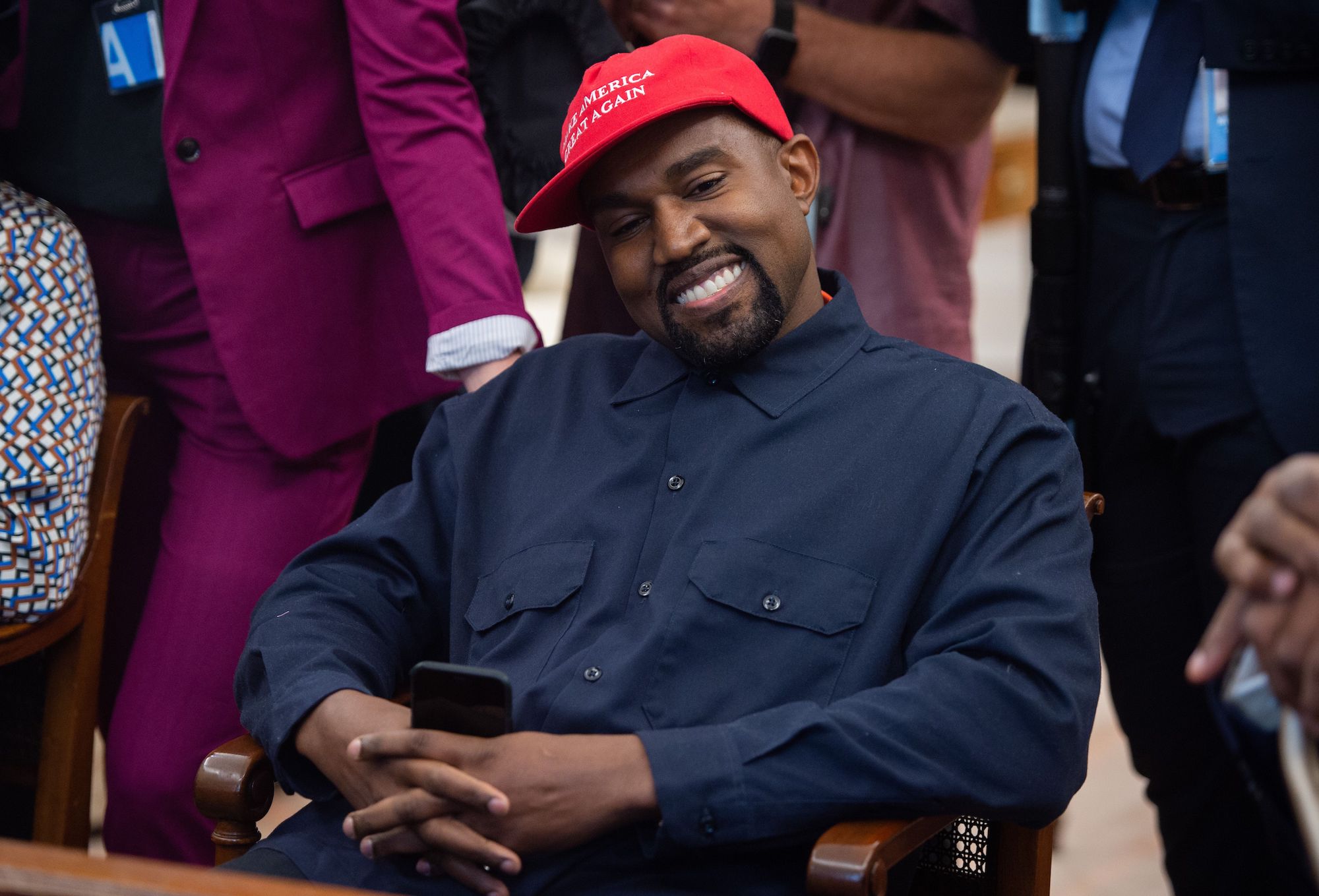 Kanye West smiling looking away from the camera wearing a red hat that says 'Make America Great Again'
