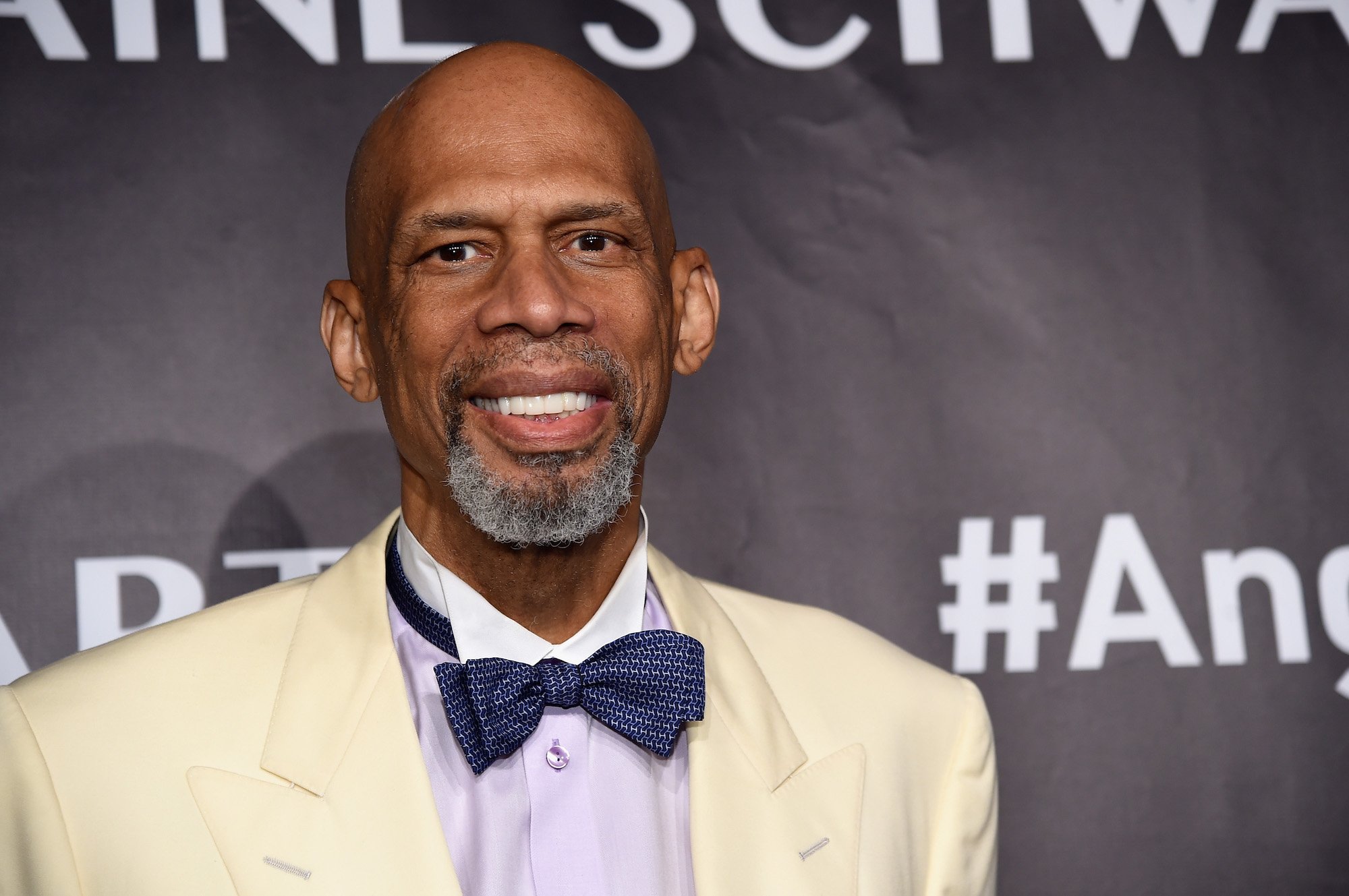Kareem Abdul-Jabbar smiling in front of a black background wearing a white suit and blue bowtie