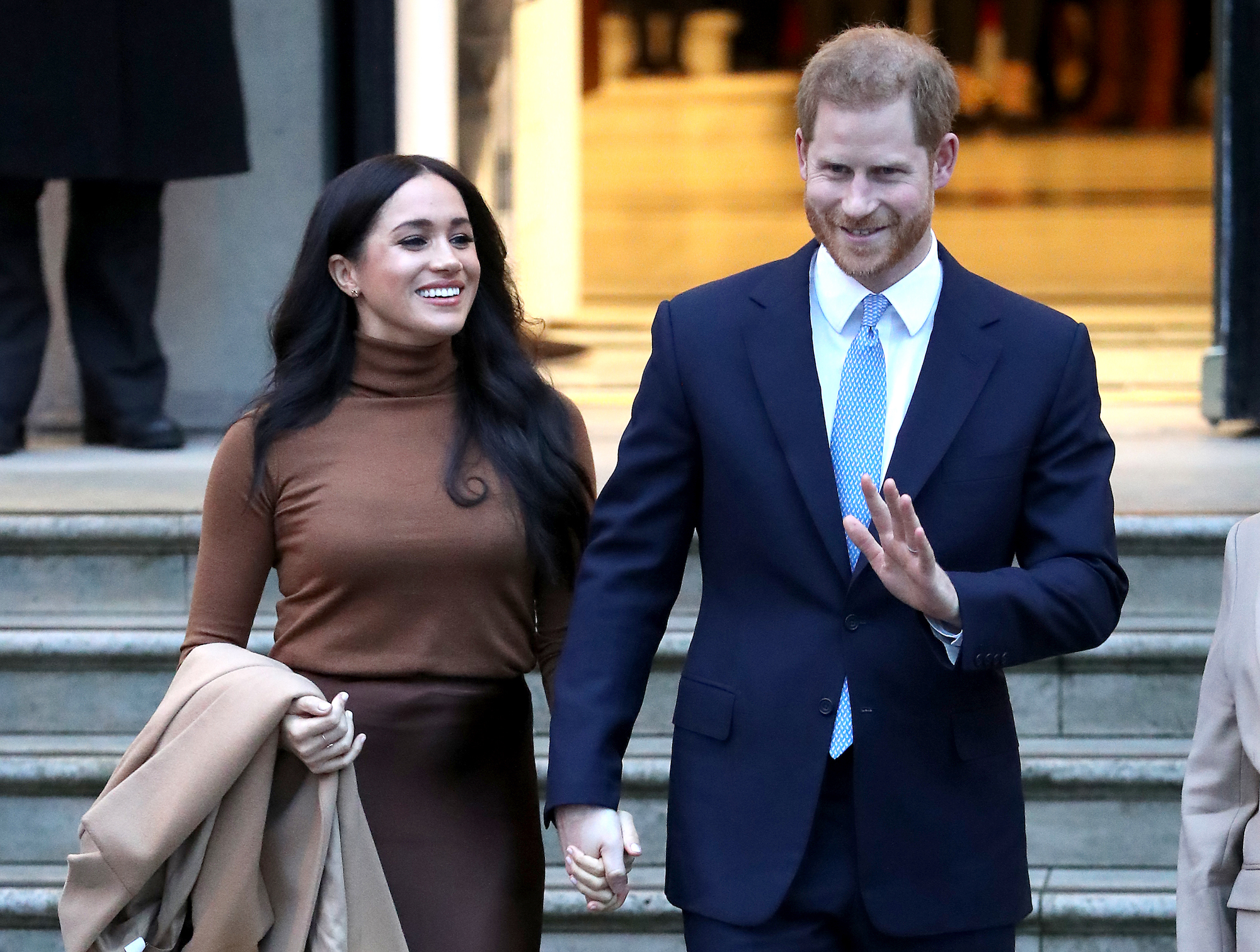 Meghan Markle smiling, looking to the side, holding hands with Prince Harry