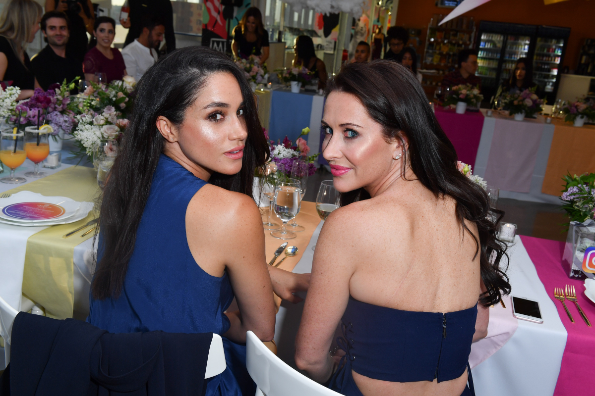 Meghan Markle and Jessica Mulroney sitting at a dining table and looking behind them towards the camera