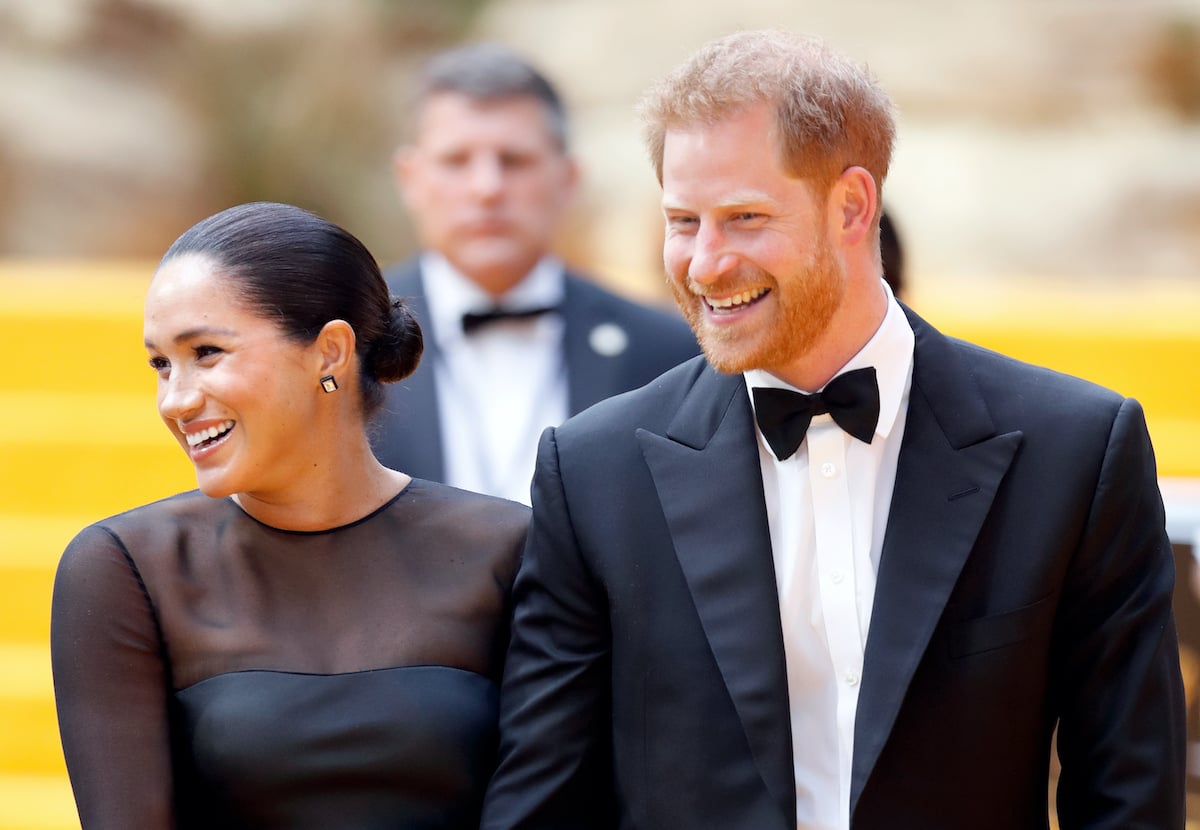 Meghan Markle wears a black dress and walks with Prince Harry in a black tuxedo as they both look to the side and smile at the premiere of The Lion King
