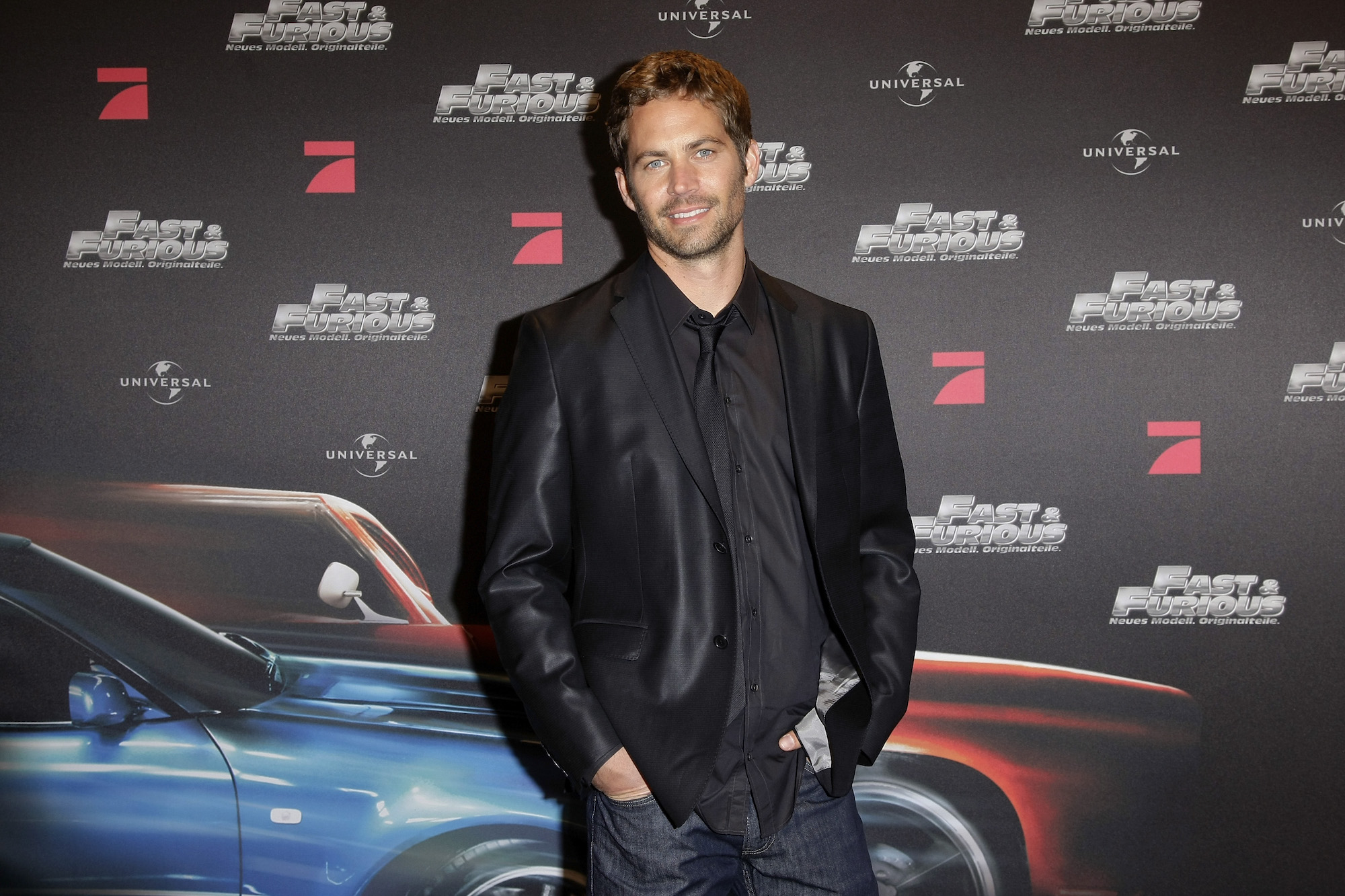 Paul Walker smiling at the camera in front of a dark background with repeating 'Fast and Furious' logos