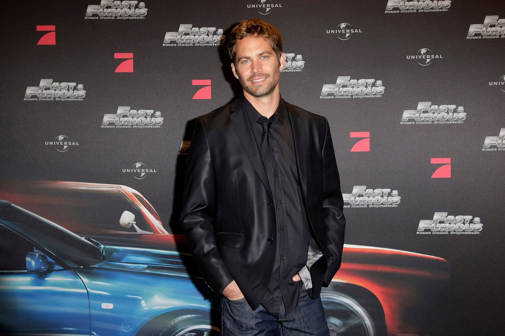 Paul Walker smiling at the camera in front of a dark background with repeating 'Fast and Furious' logos