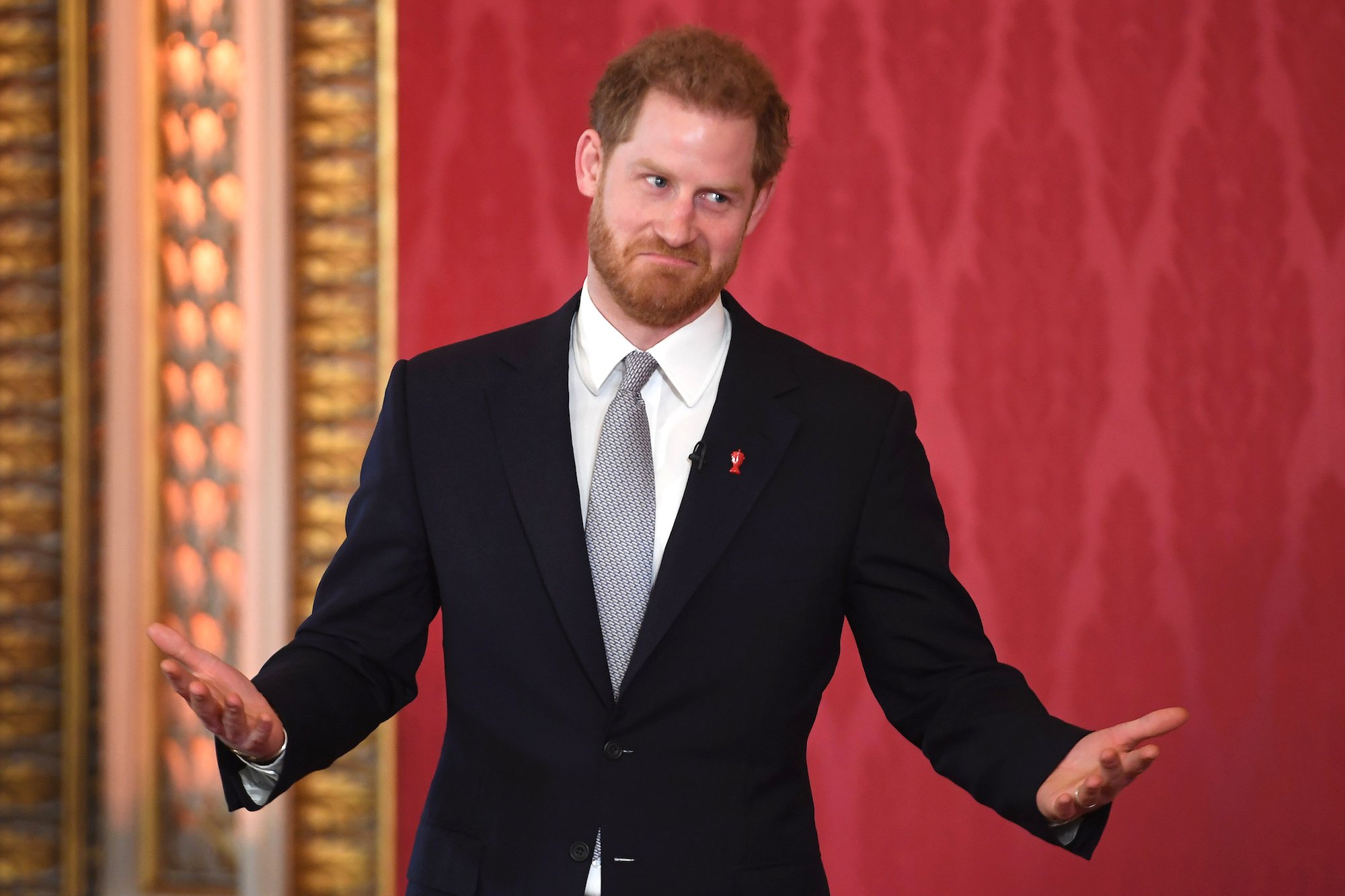 Prince Harry shrugging and smiling in front of a red background