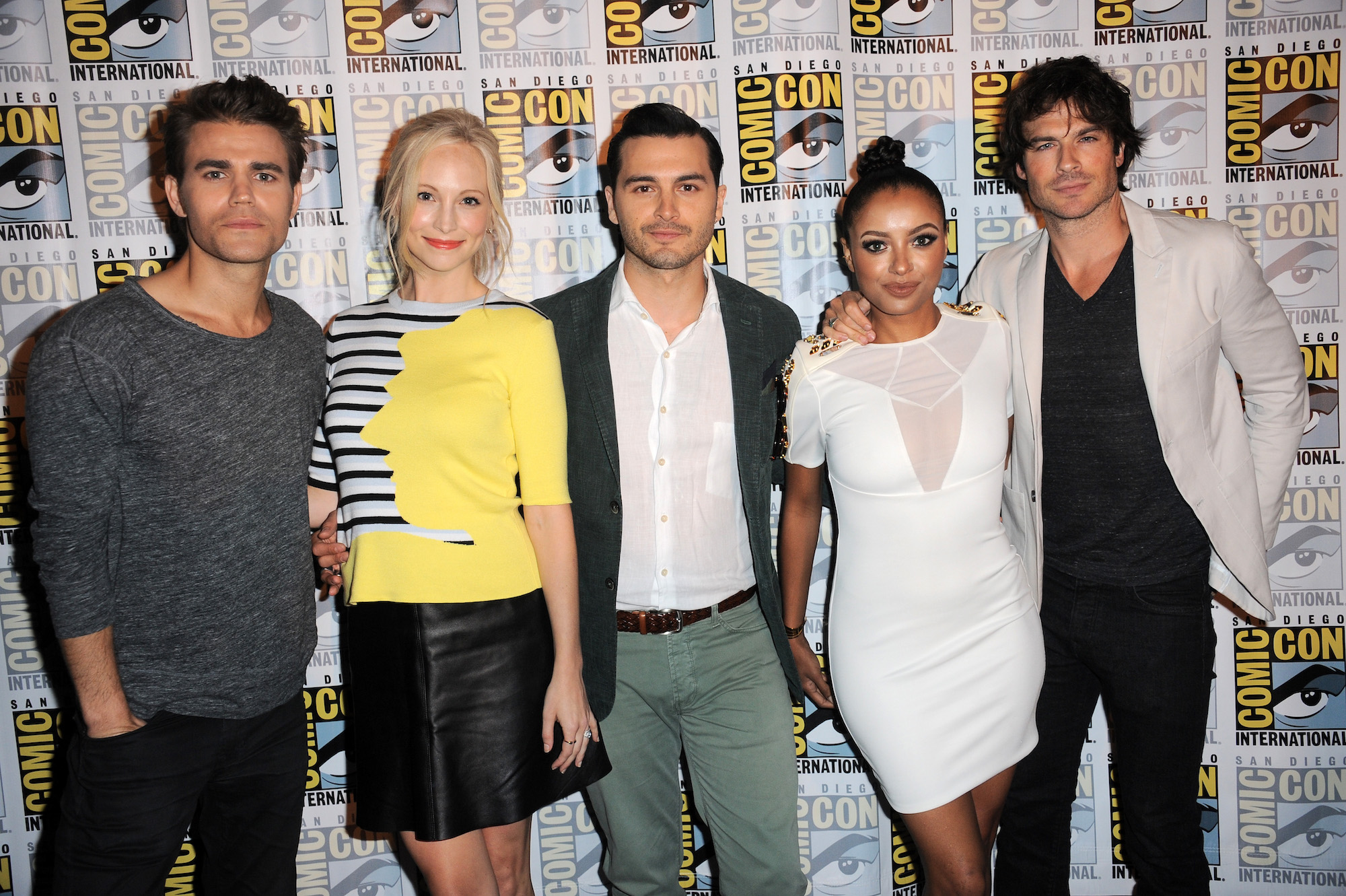 (L-R) Paul Wesley, Candice Accola, Michael Malarkey, Kat Graham and Ian Somerhalder smiling in front of a Comic Con backdrop