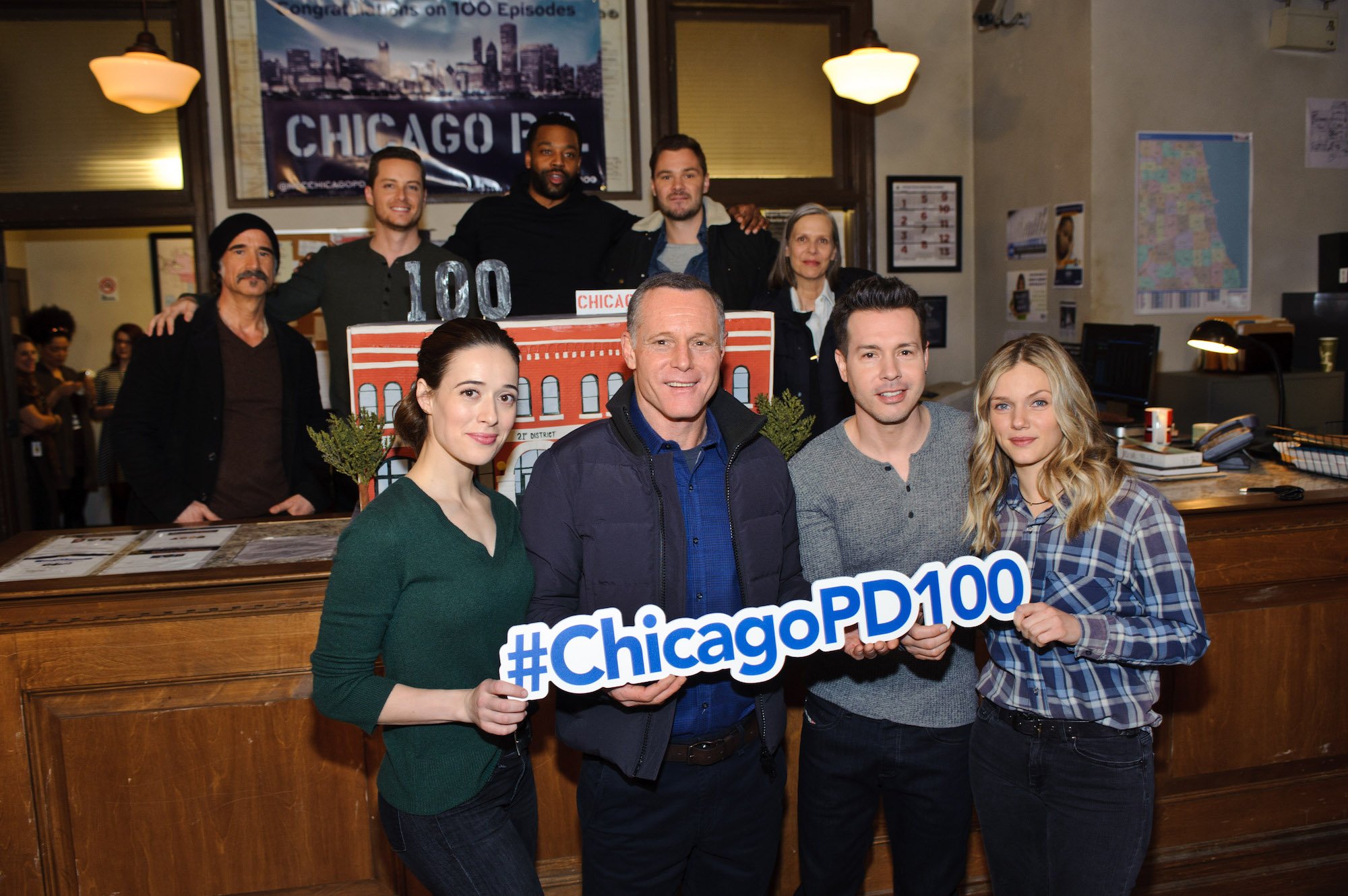 Marina Squerciati, Jason Beghe, Jon Seda,Tracy Spiridakos Elias Koteas, Jesse Lee Soffer, LaRoyce Hawkins, Patrick John Flueger, and Amy Morton smiling with a Chicago PD 100 episode prop