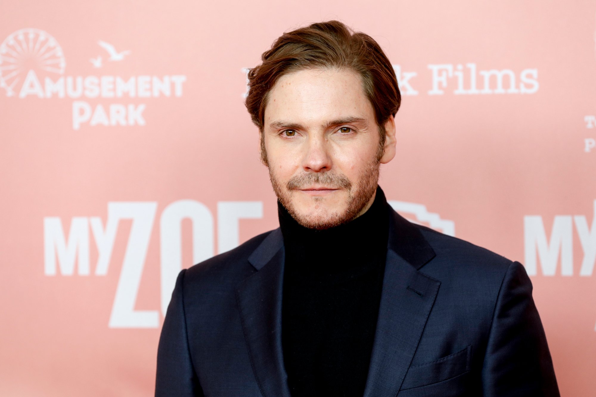 Daniel Bruehl smiling slightly in front of a pink background