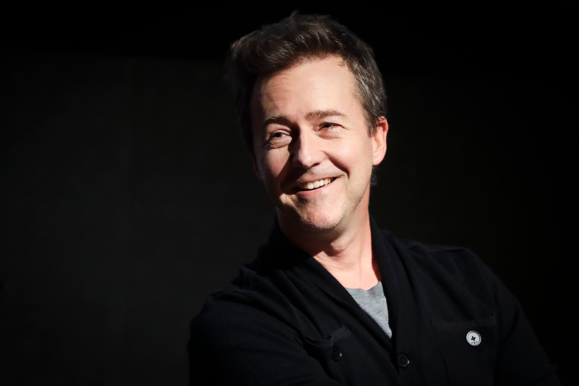 Edward Norton laughing, looking off camera, in front of a black background