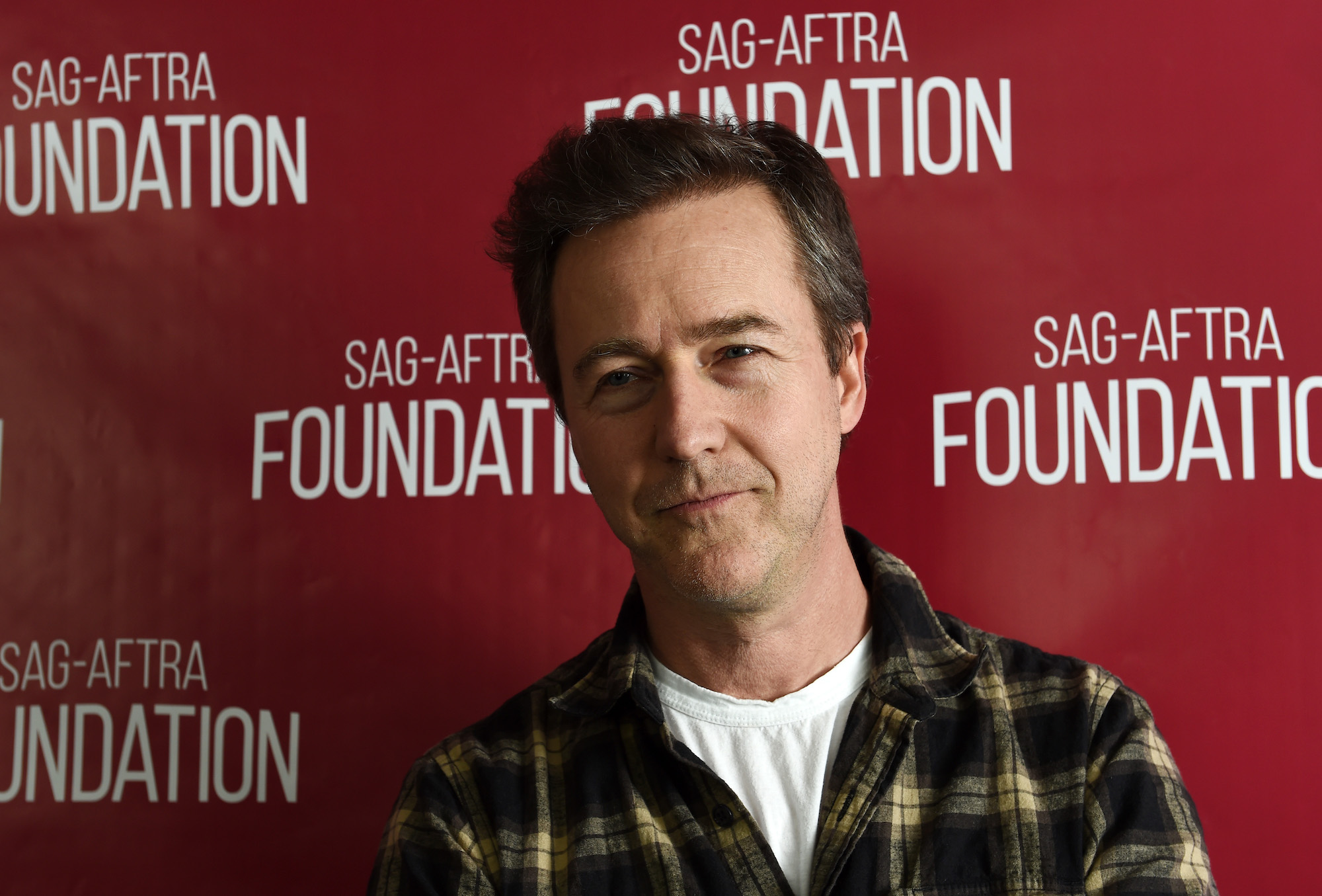 Edward Norton slightly smiling in front of a red background
