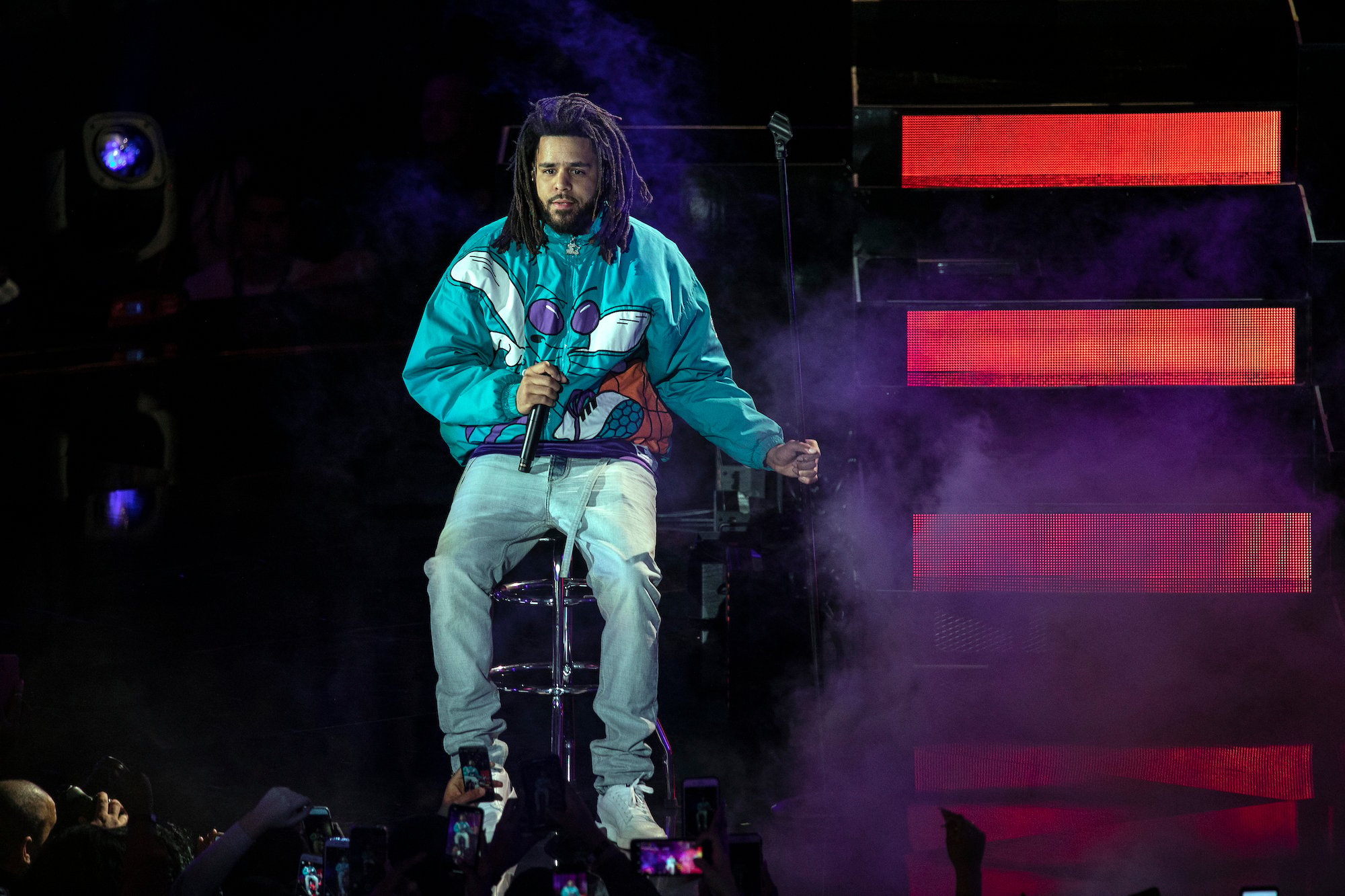 J. Cole sitting on a stool, holding a microphone on a stage