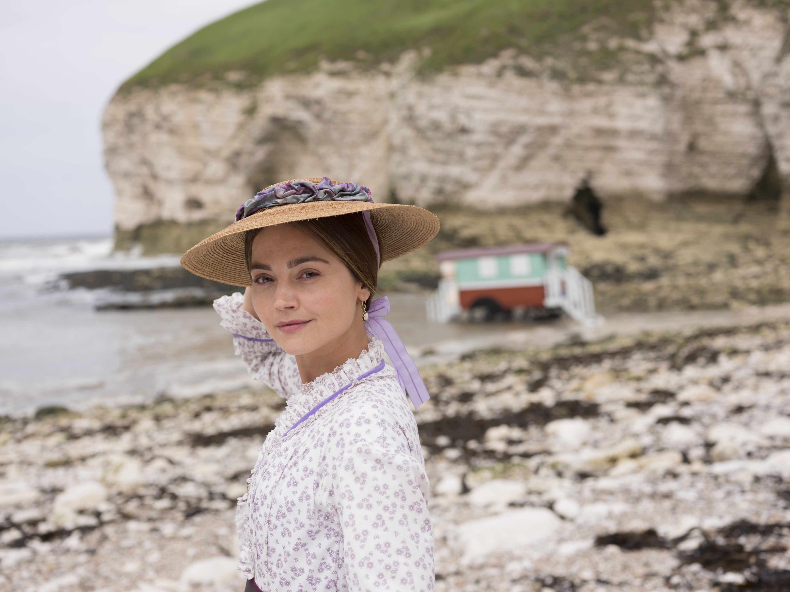 Jenna Coleman as Queen Victoria wearing a straw hat and standing on a beach 