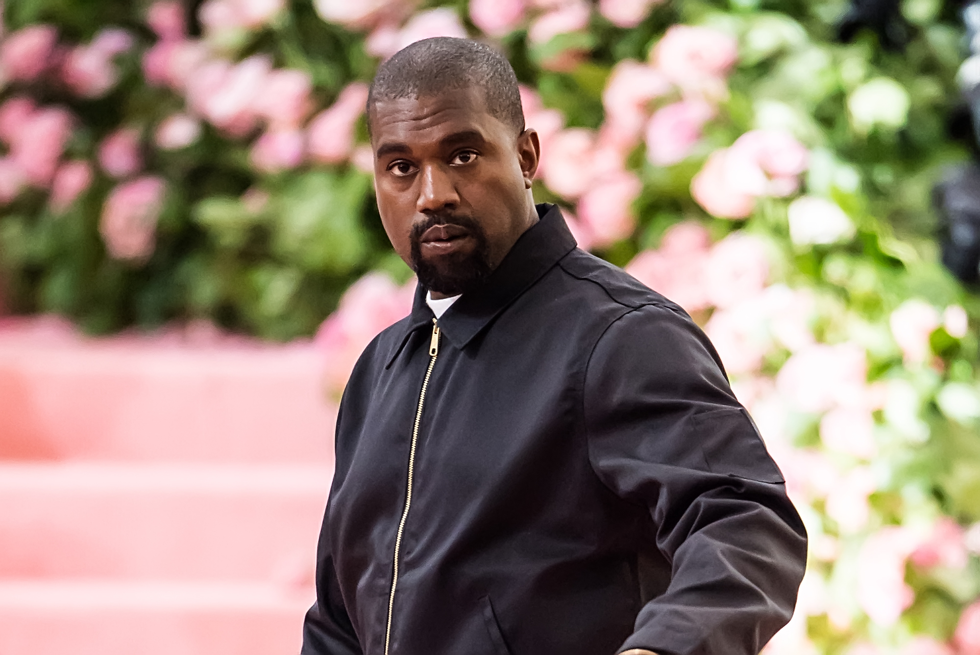 Kanye West looking at the camera, not smiling, in front of a blurry pink and green background