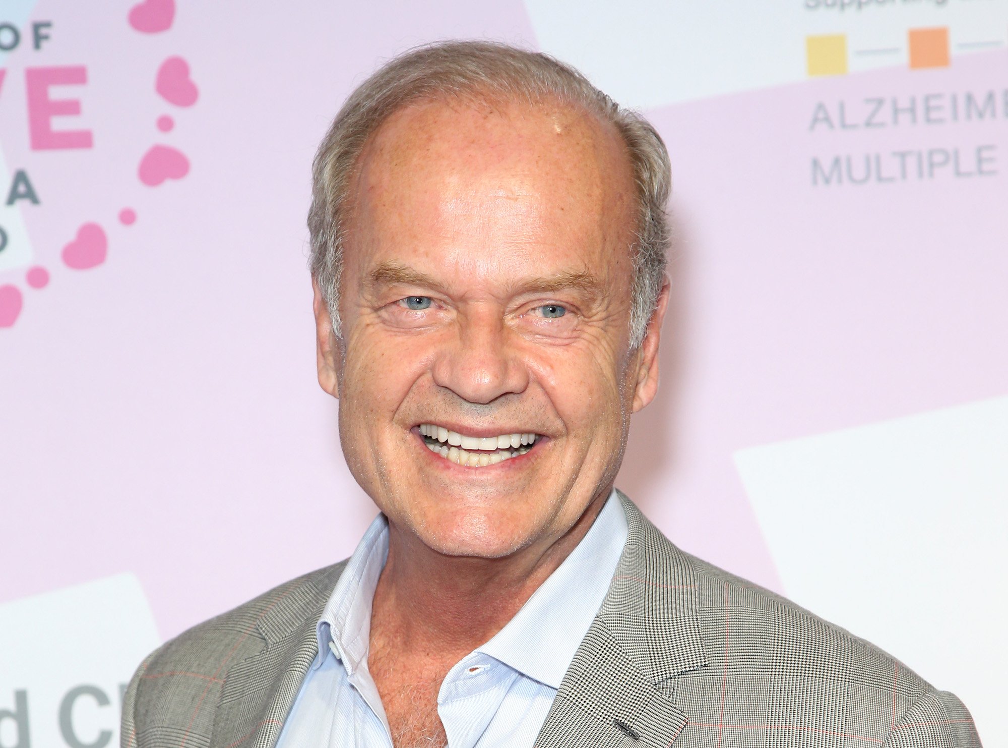 Kelsey Grammer wife one is Camille Grammer-Meyer and Kelsey Grammer spouse now is Kate Grammer. Here is Kelsey Grammer smiling in front of a pink and white backdrop