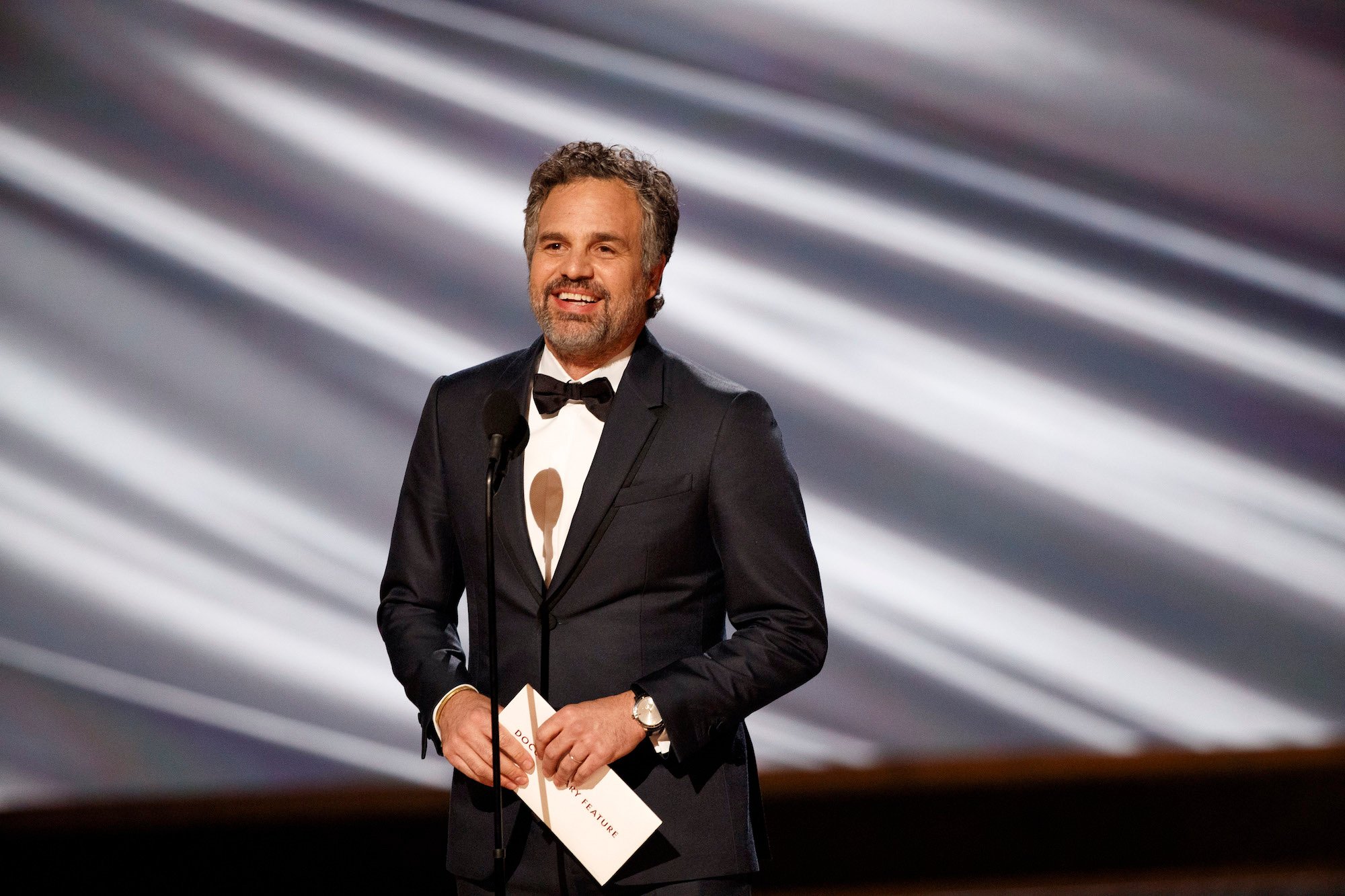Mark Ruffalo smiling in front of a gray and white background