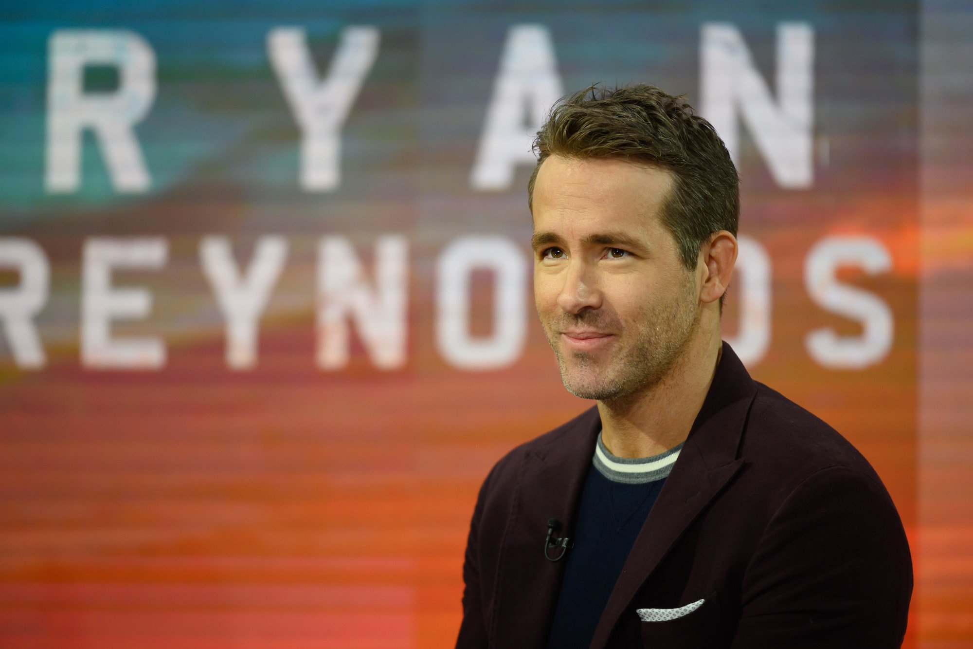 Ryan Reynolds smiling in front of an orange and blue background