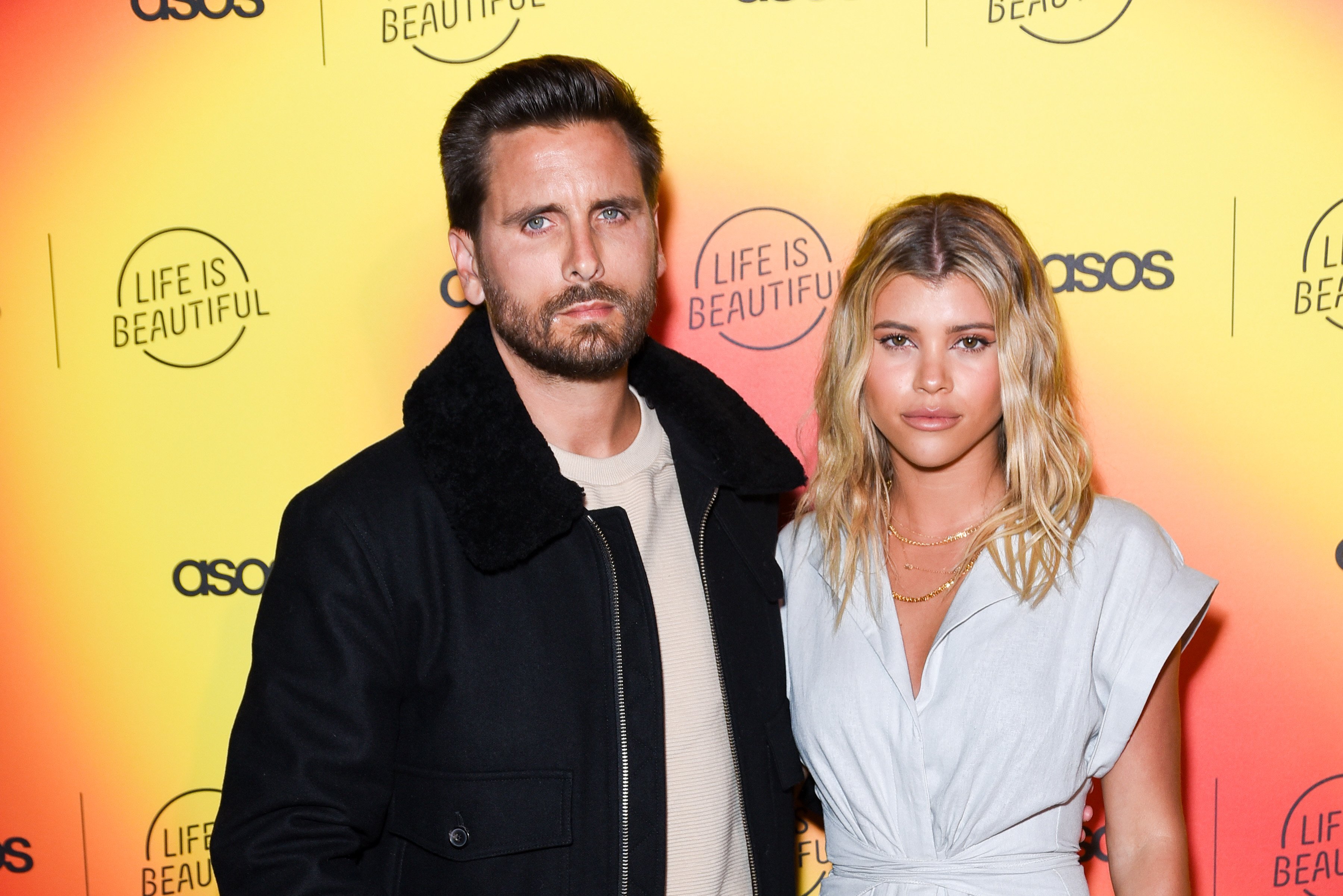 Scott Disick and Sofia Richie not smiling in front of an orange and yellow backdrop