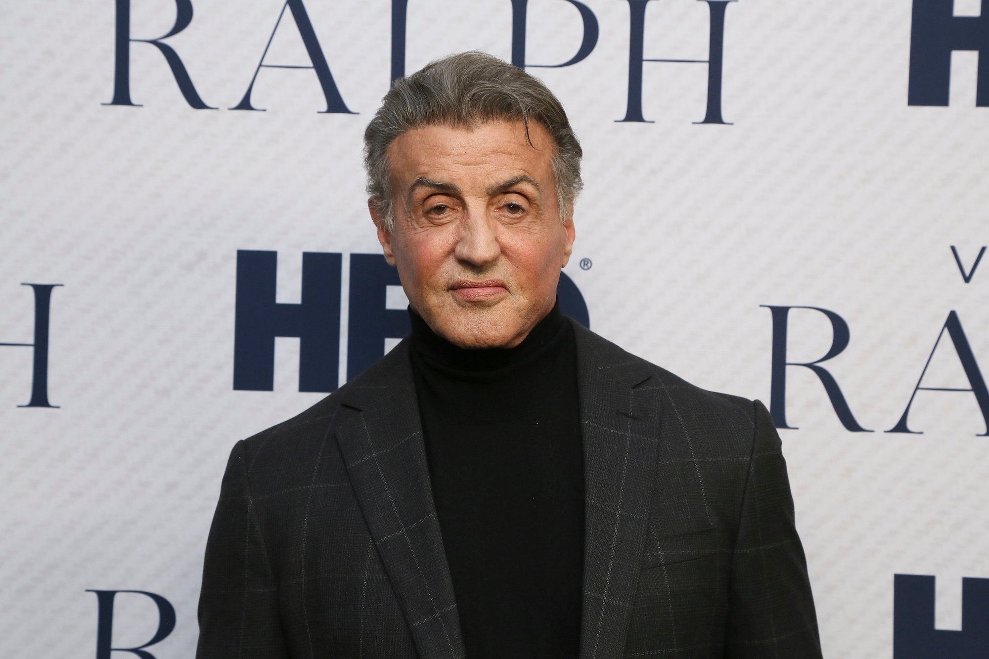 Sylvester Stallone smiling slightly in front of a white background with repeating text