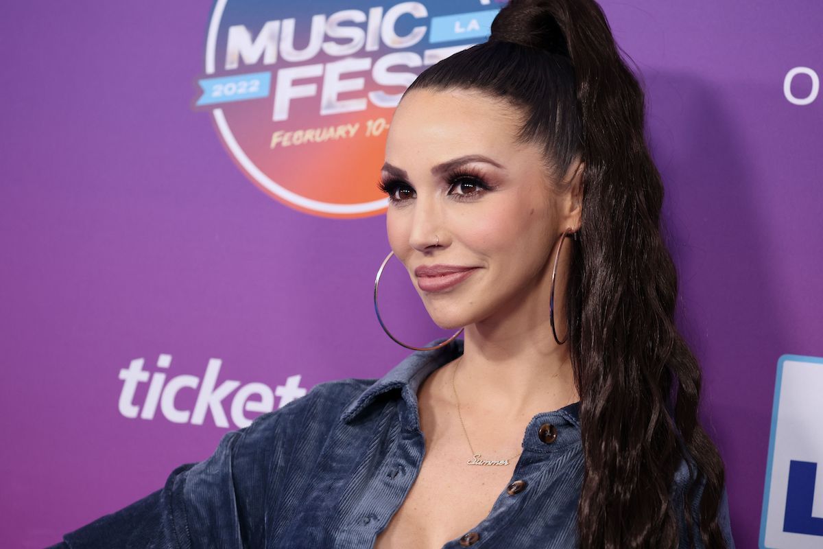 Scheana Shay, who once claimed she dated John Mayer, posing in front of a purple backdrop with her hair up in a ponytail and wearing large hoop earrings