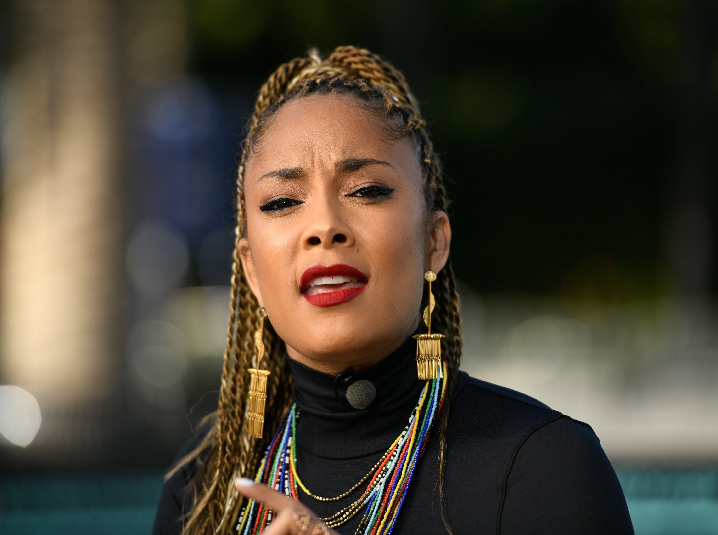Amanda Seales is photographed discussing some topic. She's wearing a black top with colorful jewelry and a braided hairstyle.